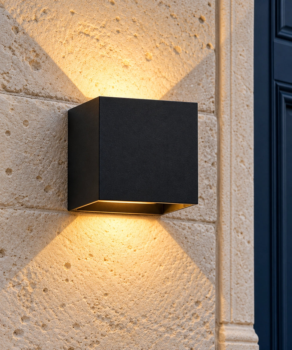 Applique extérieure cube noire allumée sur un mur en pierre, avec lumière vers le haut et le bas