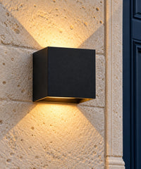 Applique extérieure cube noire allumée sur un mur en pierre, avec lumière vers le haut et le bas