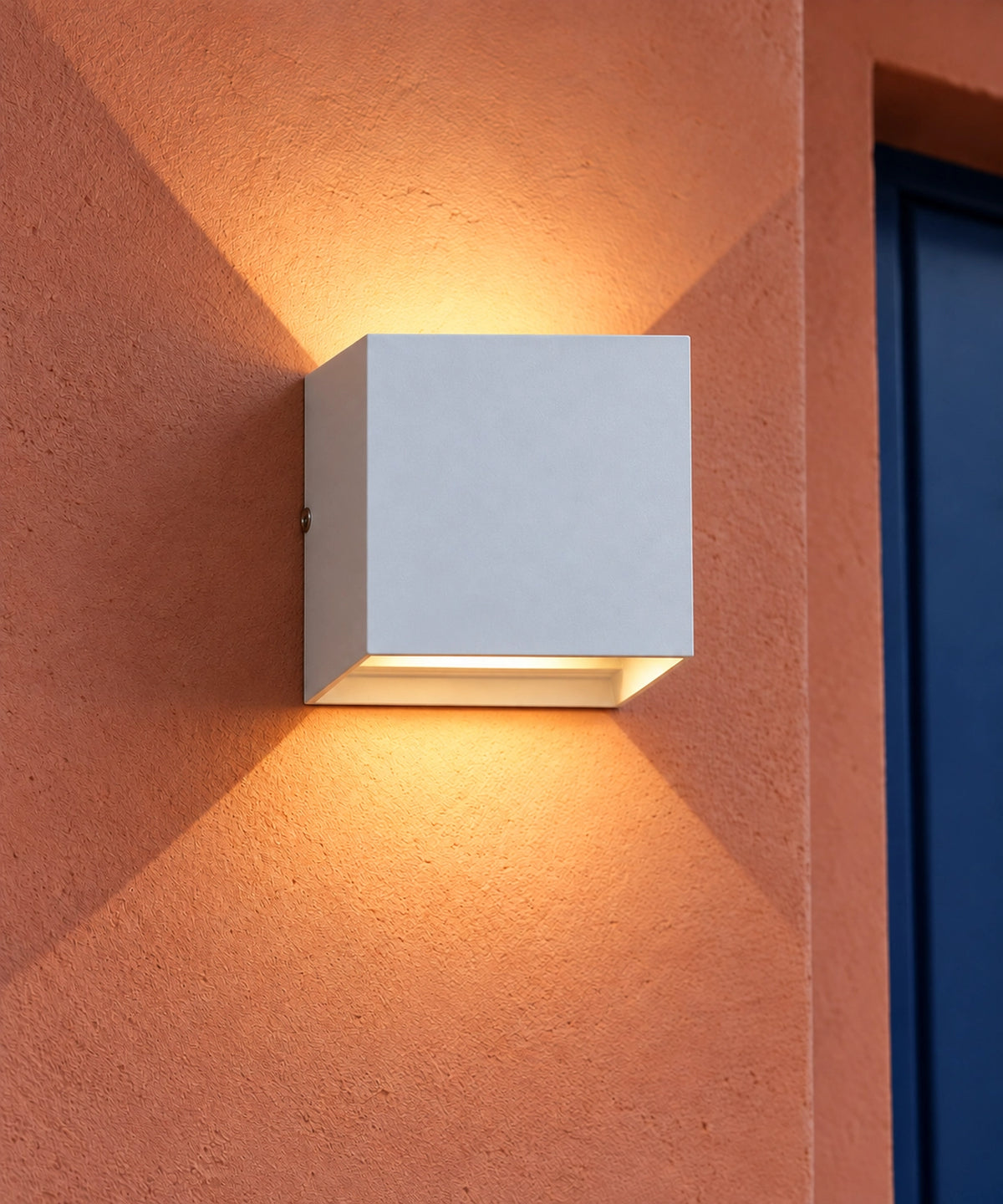 Applique cube blanche allumée sur un mur terracotta, près d’une porte sombre
