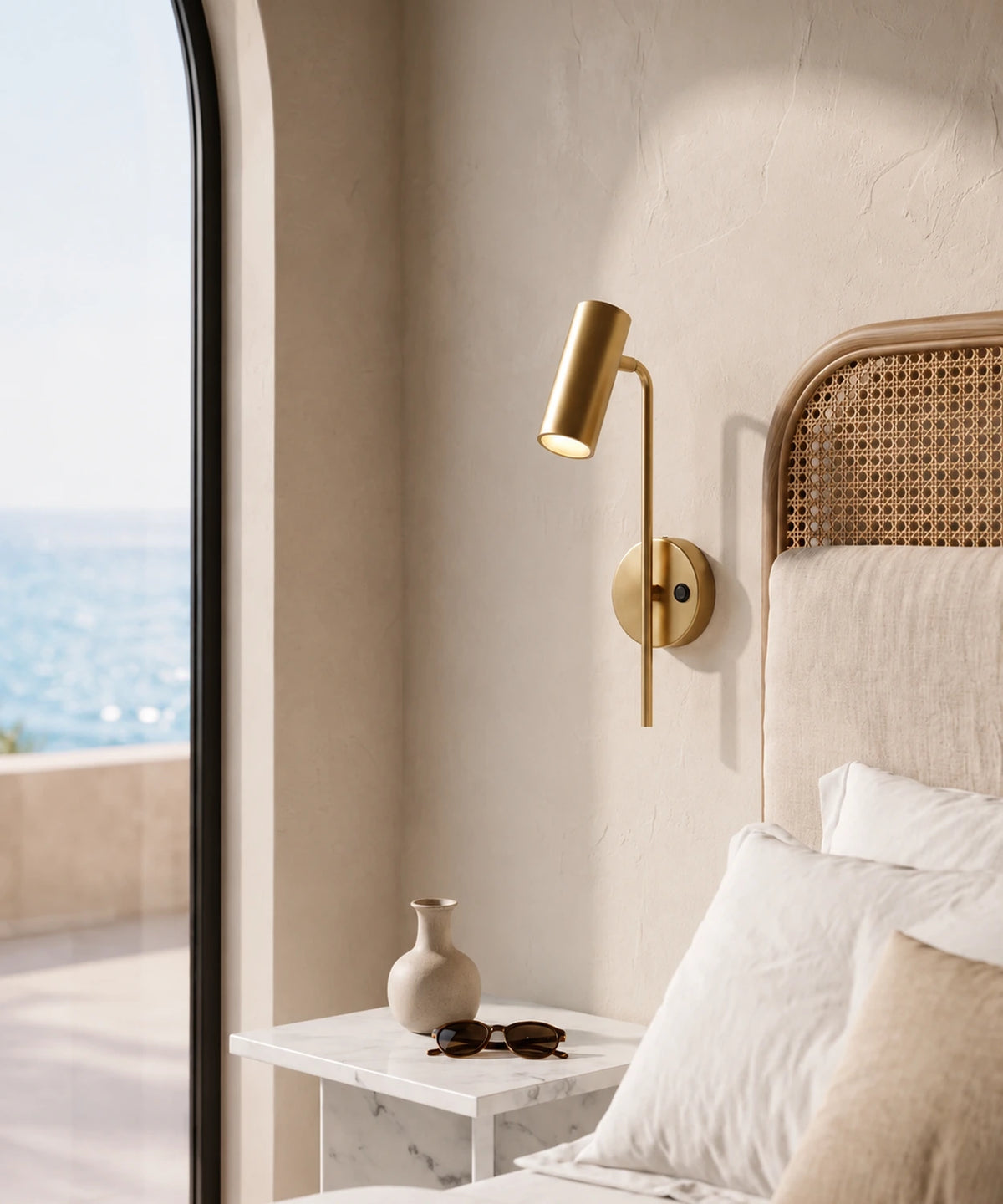 Applique liseuse orientable dorée au chevet d’un lit en rotin, fenêtre sur la mer