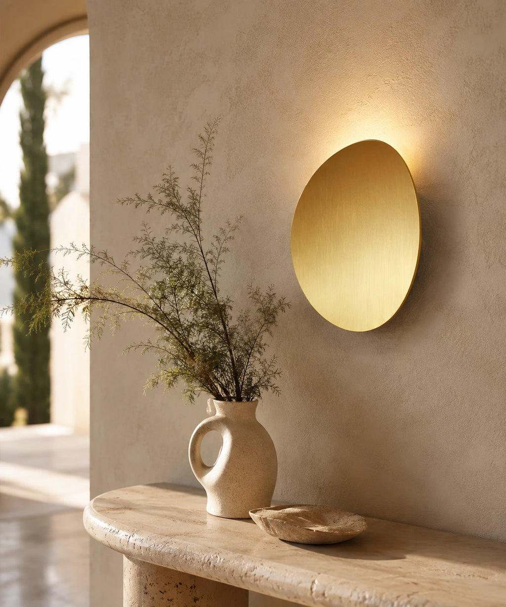 Applique murale galet or brossé au-dessus d’une console en travertin, cyprès en fond