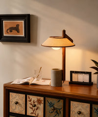 Lampe à poser en béton et lin allumée sur un meuble en bois, à côté d’un livre ouvert