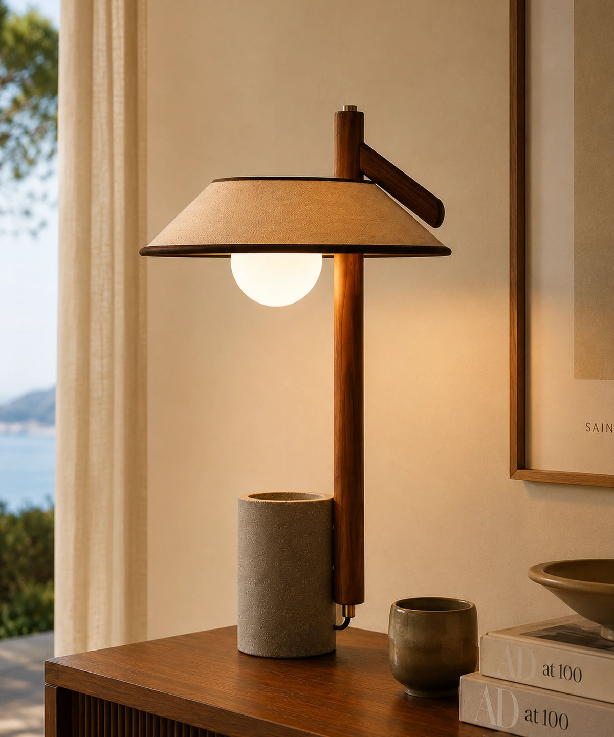 Lampe à poser béton et bois allumée sur une commode, globe opalin sous abat-jour en lin, vue sur la mer