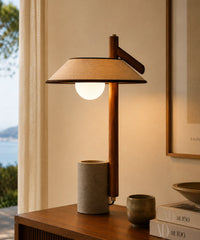 Lampe à poser béton et bois allumée sur une commode, globe opalin sous abat-jour en lin, vue sur la mer