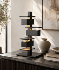 Lampe à poser design noire à caissons empilés allumée sur une console, lumière rasante sur les plateaux