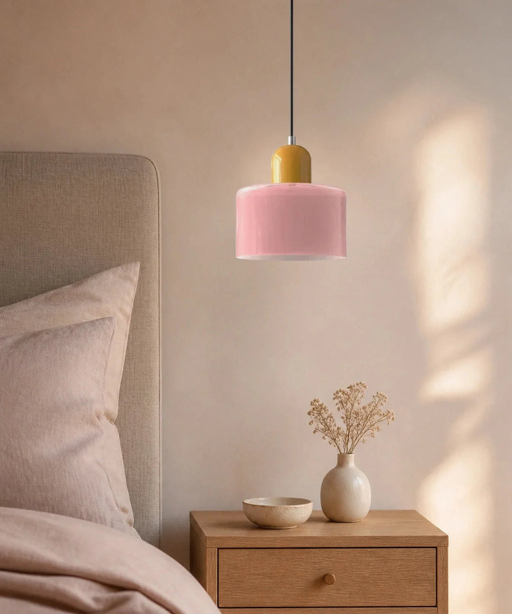Suspension cylindre en verre rose et culot jaune moutarde au-dessus d’une table de chevet